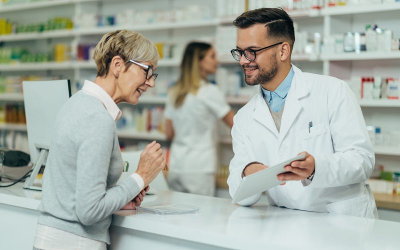 Young male pharmacist giving prescription medications to senior female customer in a pharmacy with female pharmacist in the background
